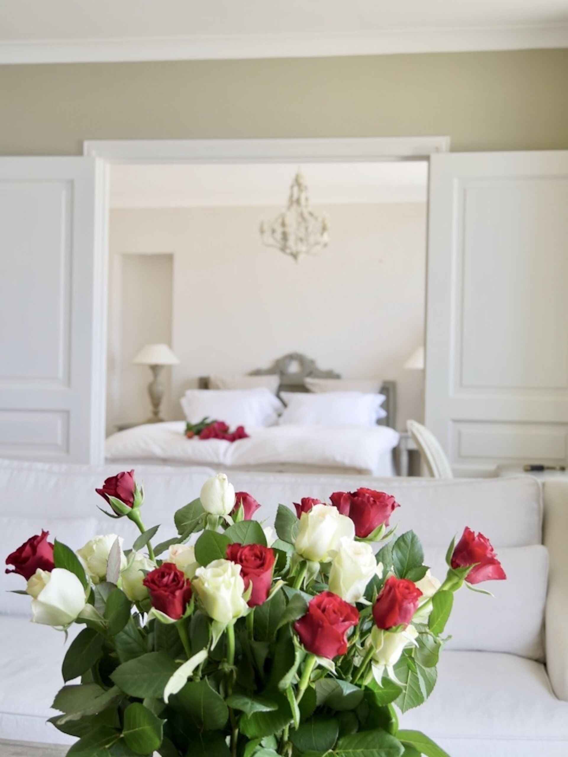 A bedroom with a bed, a chandelier, and a vase of roses in the foreground.