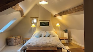A cozy attic bedroom with a sloped ceiling, a bed with floral bedding, two bedside lamps, a small window, and a rustic wooden beam. - Cottage and private garden - rural bliss for two (or 4 with mezzanine) (Corsaint)