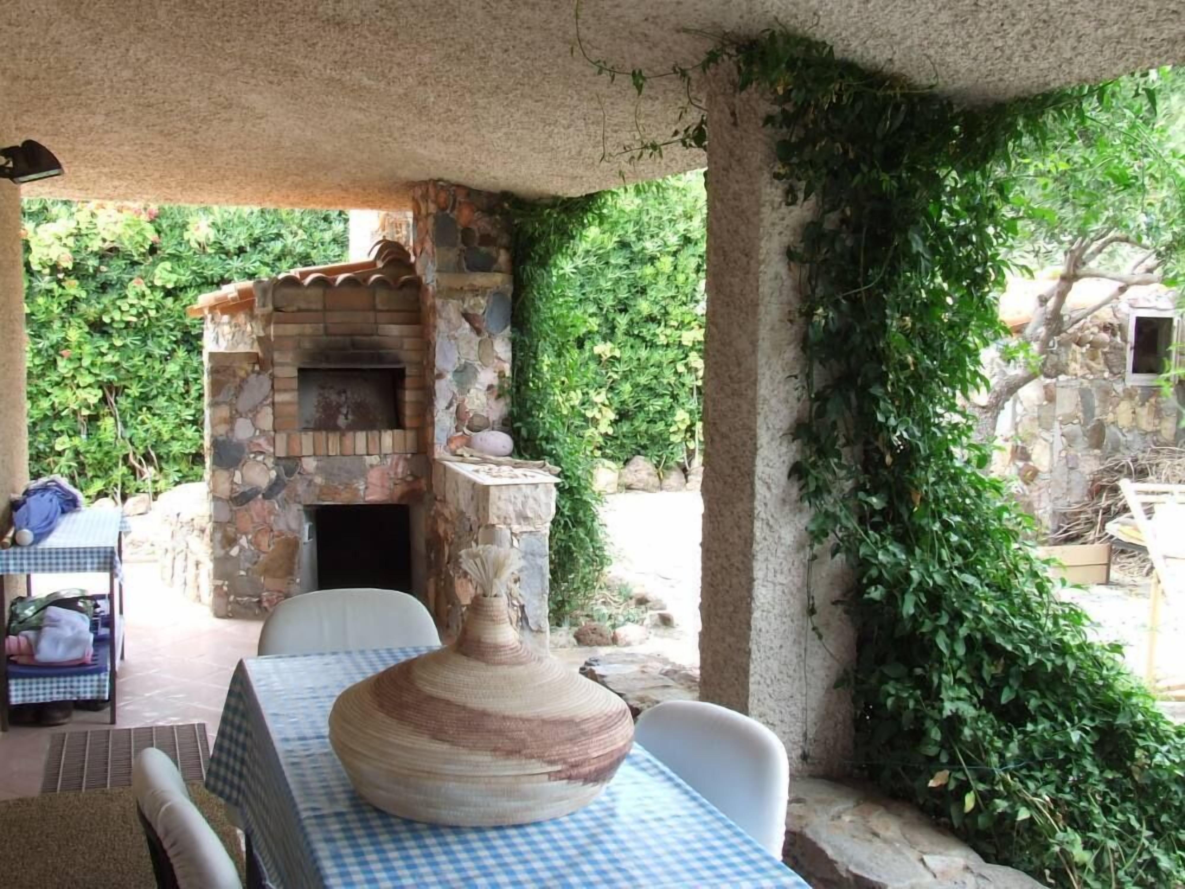 Une terrasse extérieure avec un espace barbecue en pierre, une table recouverte d’une nappe à carreaux et un grand pot en céramique.