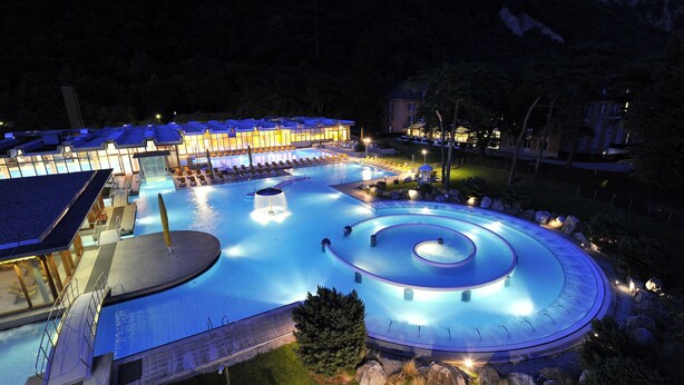 Une grande piscine extérieure dotée d’un élément décoratif aquatique circulaire, entourée de chaises longues et bordée d’un bâtiment dont les fenêtres sont illuminées.