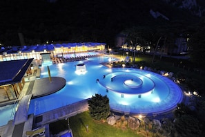 A large outdoor swimming pool with a circular water feature, surrounded by lounge chairs and a building with illuminated windows. - Historic chalet from 1714 in Plans-sur-Bex, Gryon-Villars-Les Diablerets (Les Plans-sur-Bex)