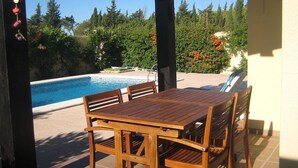 Een houten tafel en stoelen staan onder een pergola, met uitzicht op een zwembad en een omgeving van bomen en struiken.