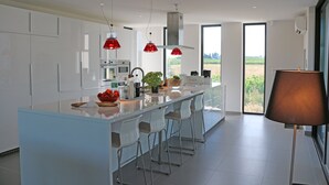 A modern kitchen with white cabinets, a large island with bar stools, red pendant lights, and a view of the countryside through large windows.
