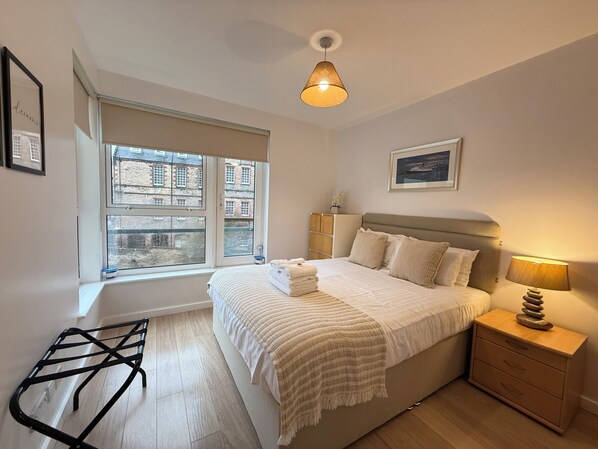 A bedroom with a bed, bedside table, lamp, and a window with a view of buildings.