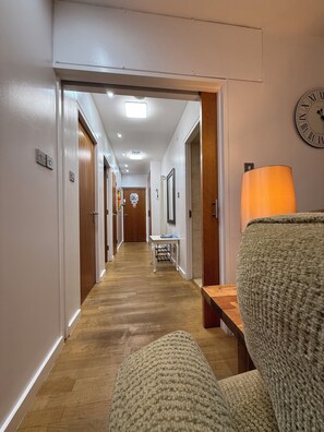 A hallway with wooden flooring, a clock on the wall, and a lamp on a table.