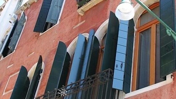 Un bâtiment rouge doté de nombreuses fenêtres, dont certaines sont pourvues de volets, et d’un balcon avec une rambarde en métal.