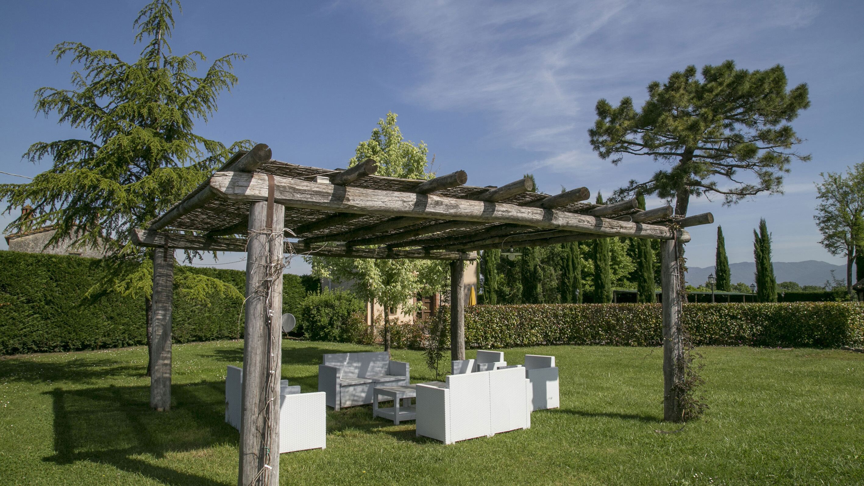 Une pergola en bois avec des meubles d’extérieur blancs, située dans un coin de jardin verdoyant.