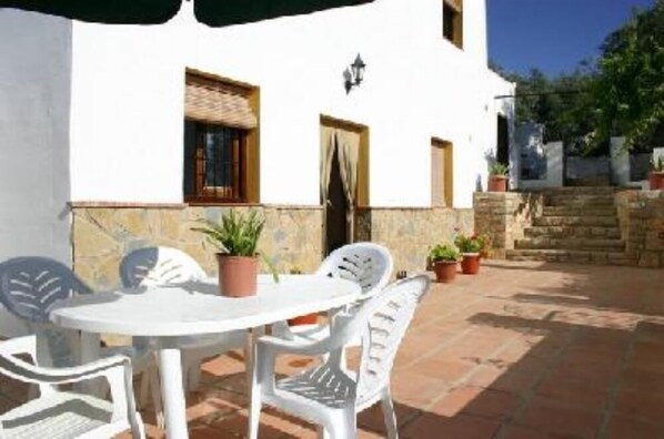 An outdoor patio with a round table and white plastic chairs, surrounded by potted plants.