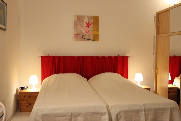 A hotel room with two beds, red curtains, a wooden nightstand with a lamp, and a painting on the wall.