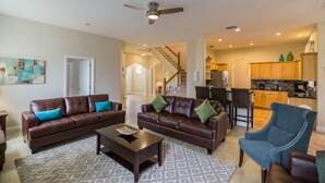 A spacious living room with a brown leather sofa, a blue armchair, and a coffee table. A staircase leads to the kitchen in the background. - 🏡 7 Bed,7 Bath | Large pool & H/Tub | Large Smart TVs & Sonos every Room I WiFi (Davenport)