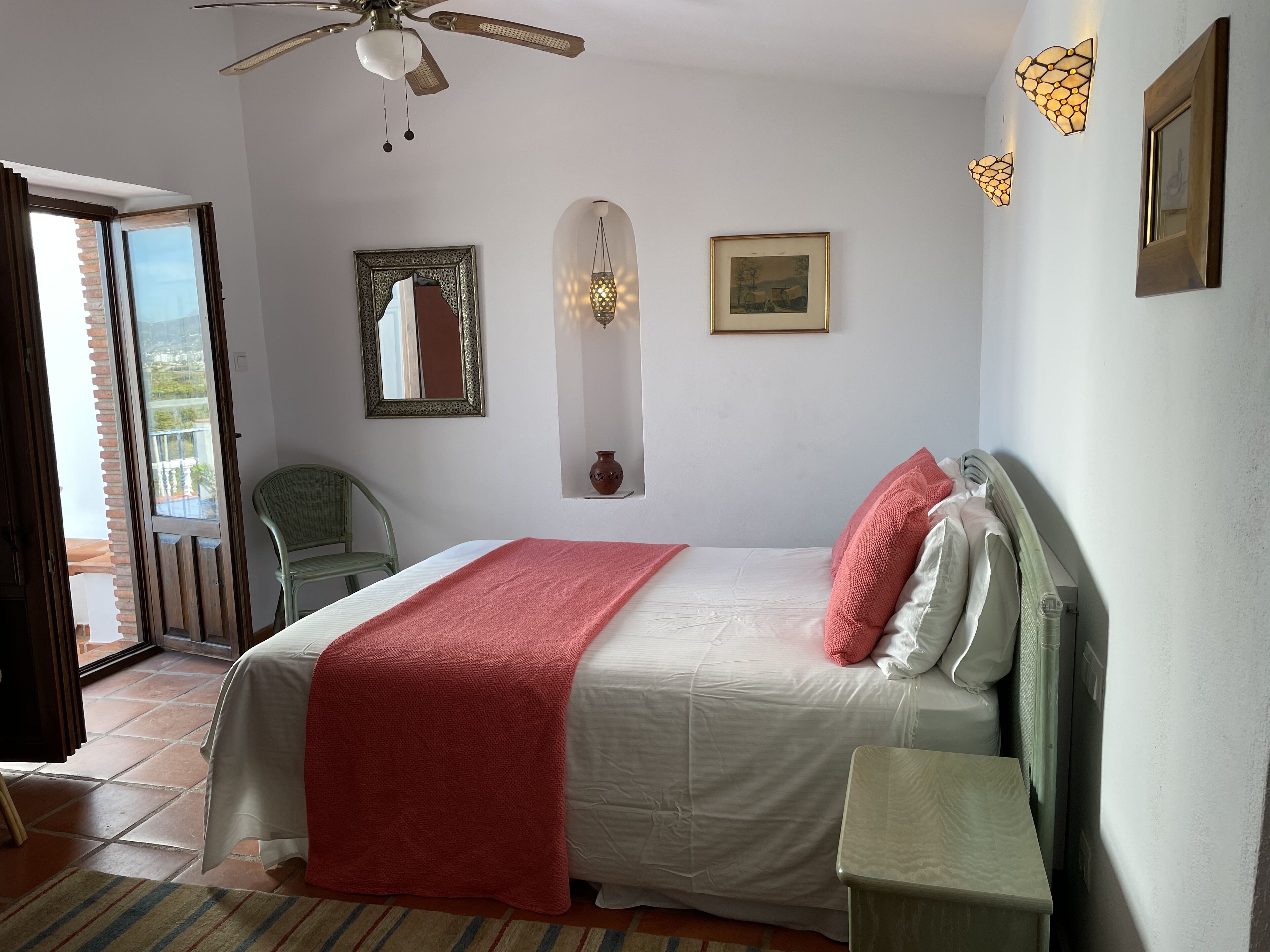 Ein Schlafzimmer mit Bett, Nachttisch, Deckenventilator, Fenster und einer Tür, die auf einen Balkon führt.