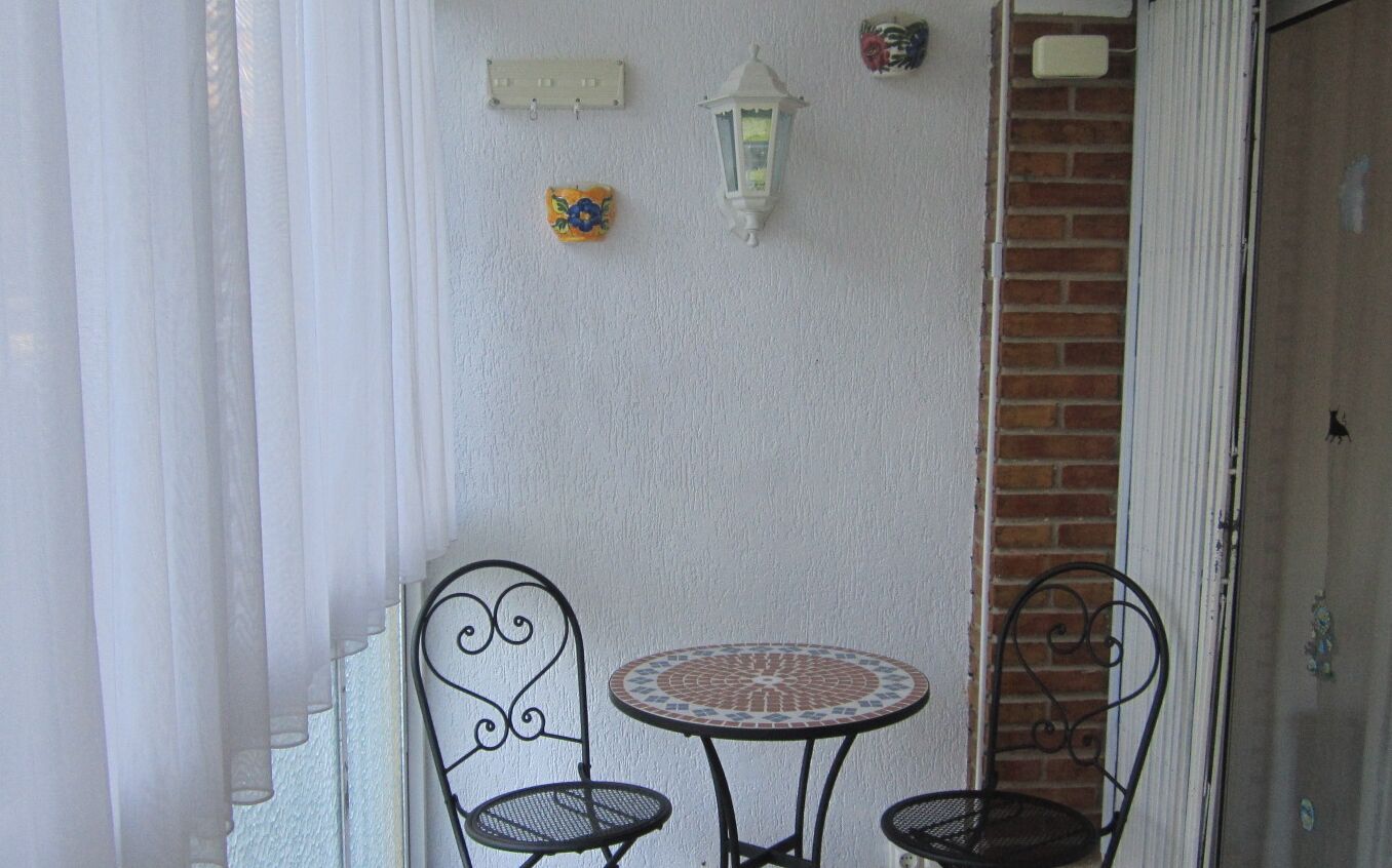 A small outdoor seating area with two metal chairs and a round table, a wall-mounted light fixture, and a decorative tile.