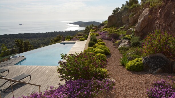 A pool with a wooden deck, surrounded by purple flowers and rocks, overlooking a coastal view. - Villa with breathtaking views of Pinarello bay and beach (Zonza)