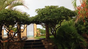A stone staircase leading to a gate, surrounded by lush greenery and a stone building with a view of the sea.