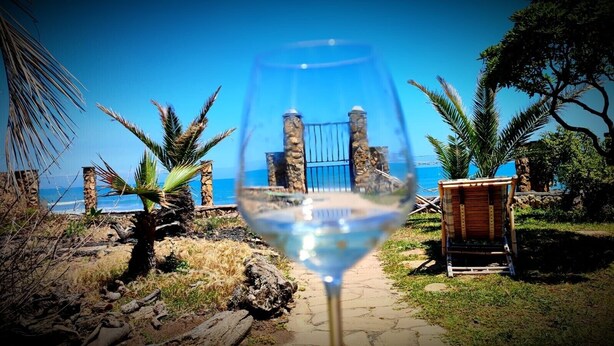 A glass of white wine with a clear view of a beach and a stone structure in the background.