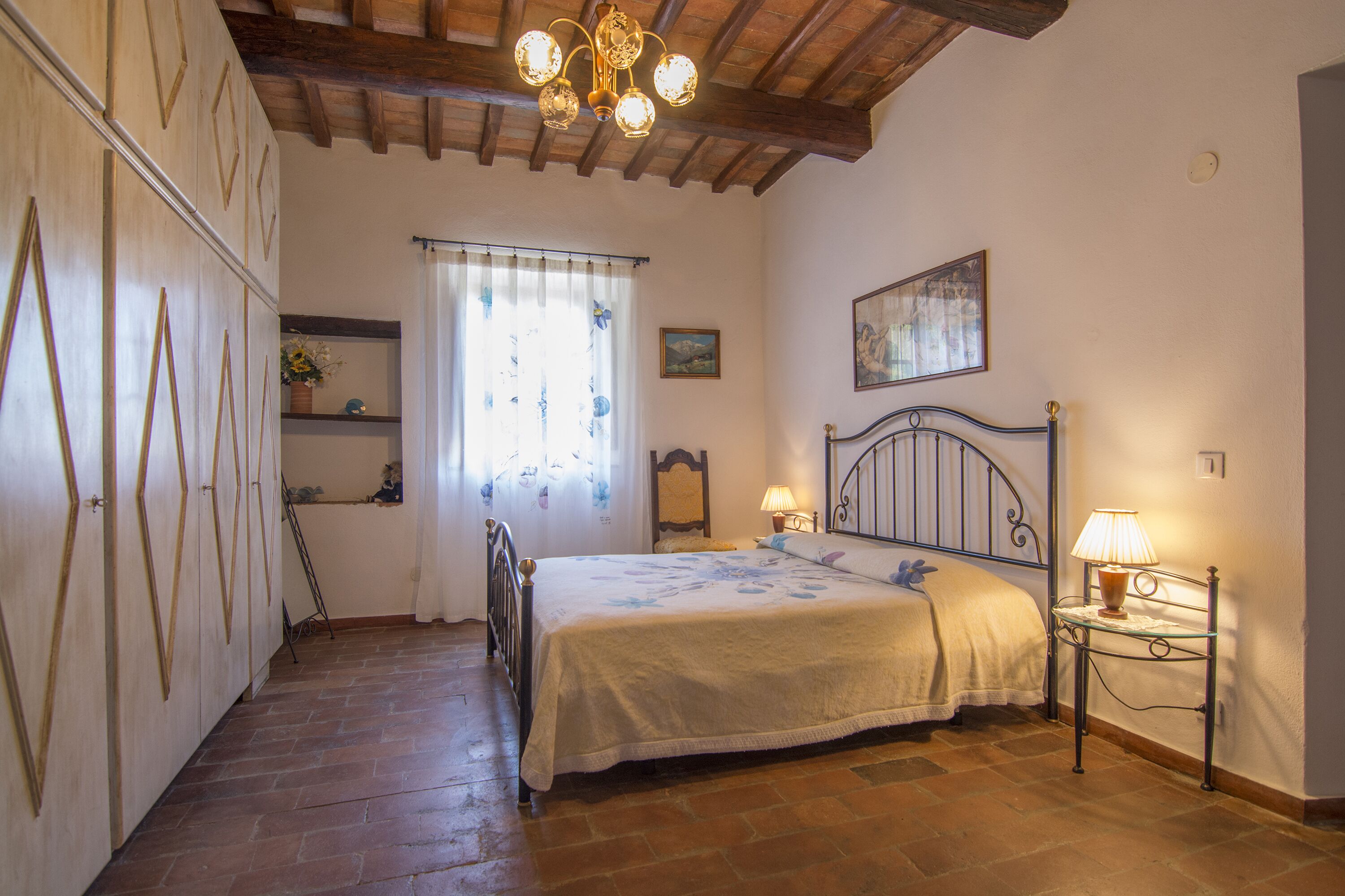 A bedroom with a bed, a bedside table with a lamp, a window with curtains, a wooden wardrobe, and a painting on the wall.