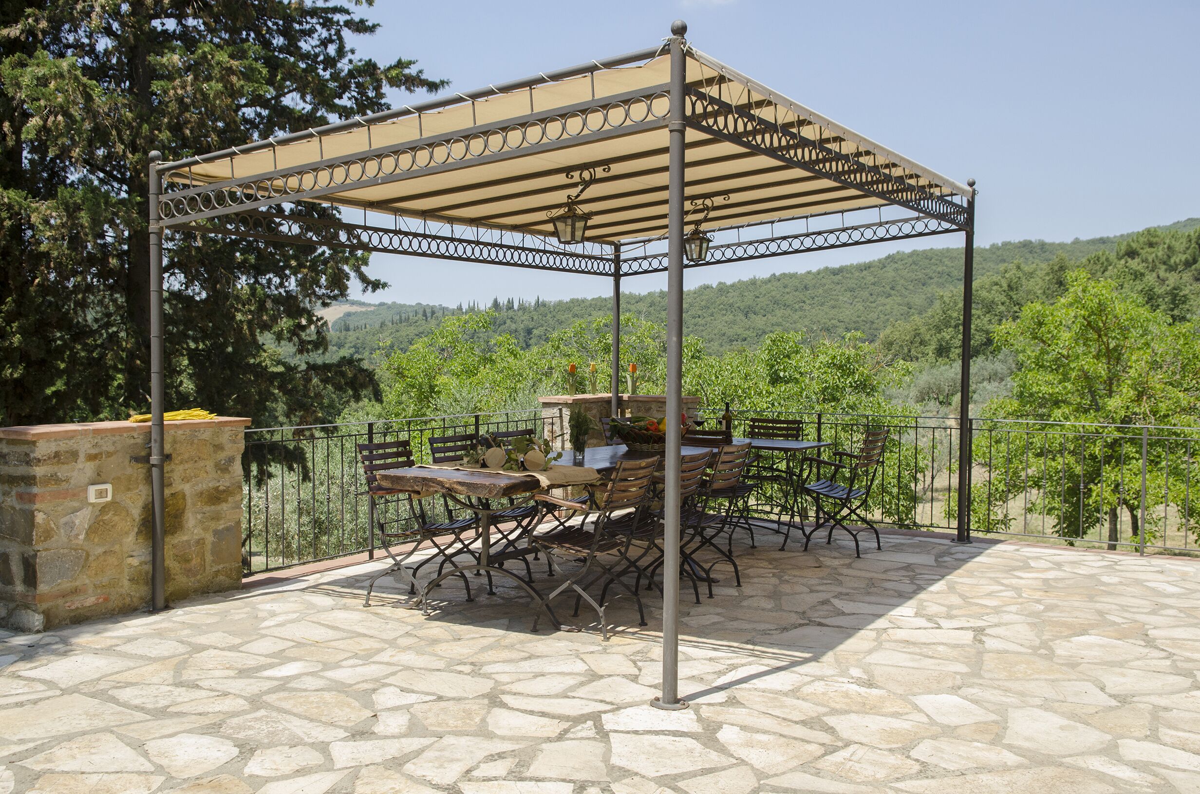 Um pátio externo com piso de pedra, uma pérgola com cobertura bege e mesas e cadeiras de estrutura metálica.
