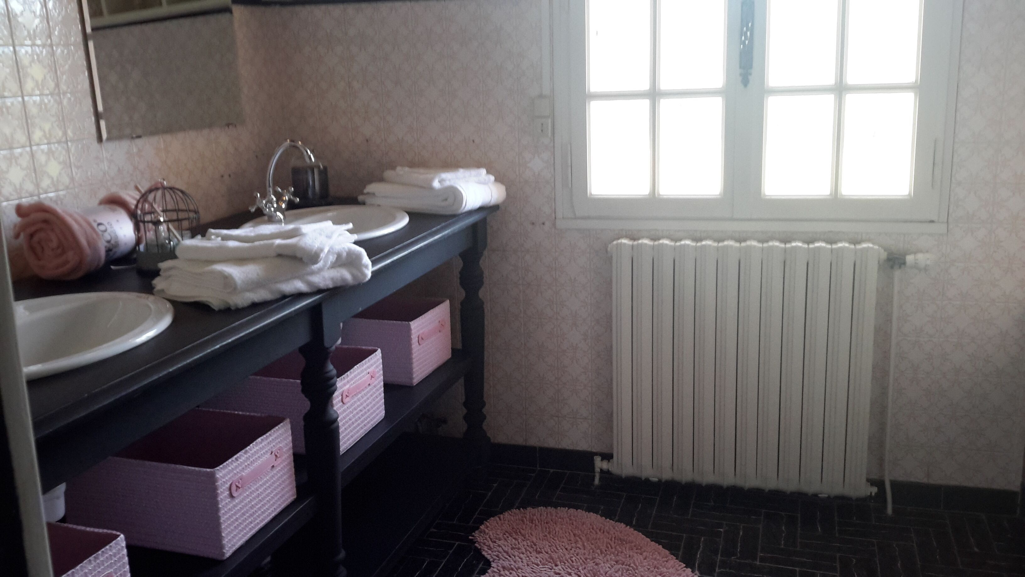 Une salle de bain équipée d’un lavabo, d’un miroir et d’un porte-serviettes. Il y a une fenêtre avec des stores et un radiateur.