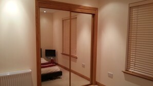 Une chambre avec un miroir coulissant intégré dans une porte, un lit, une télévision et une fenêtre avec des stores.