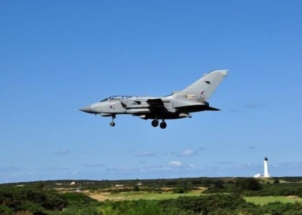 Un avión militar volando, con un faro al fondo.