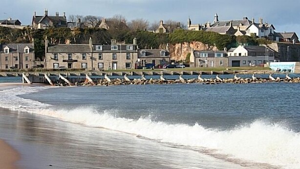 Une ville côtière avec des bâtiments en pierre, une plage de sable et une digue rocheuse où se perchent des mouettes.