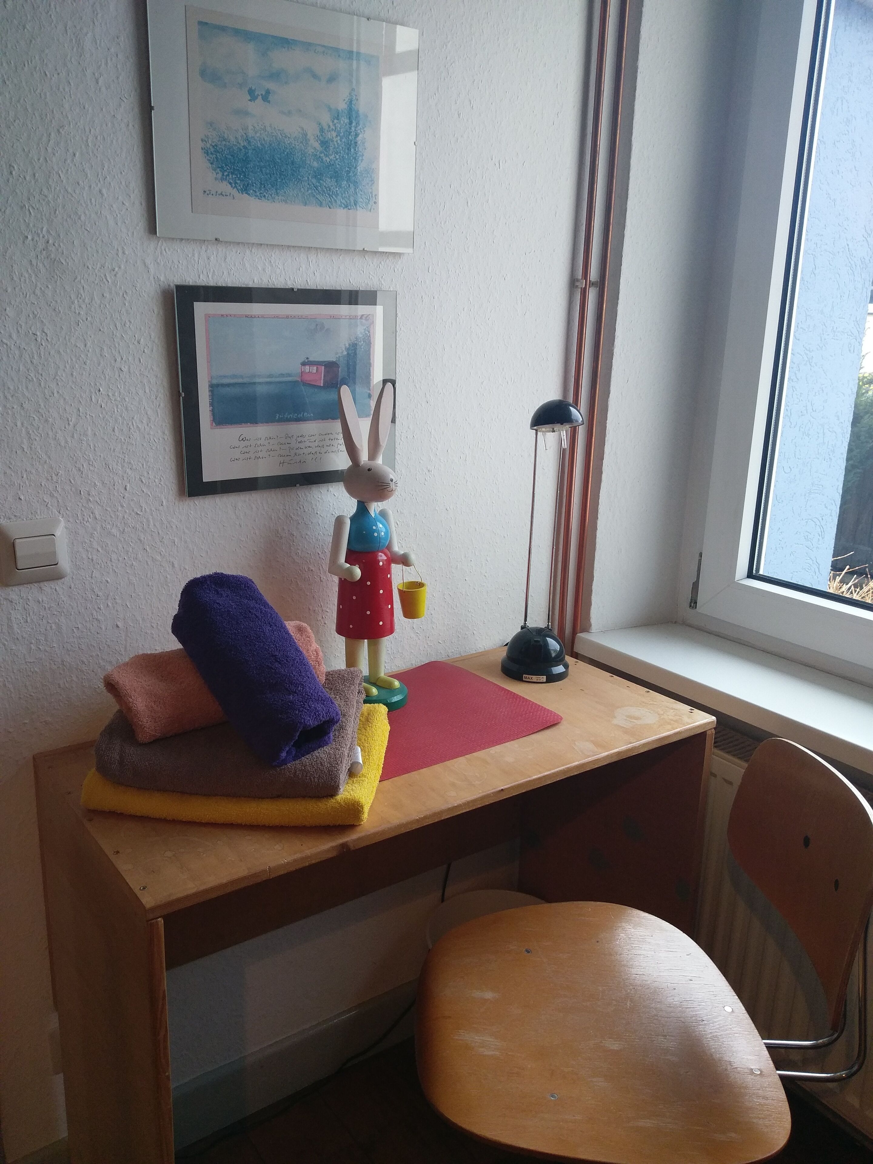 A small, neatly organized room with a wooden desk, a chair, a framed picture on the wall, and a window with a view of trees.