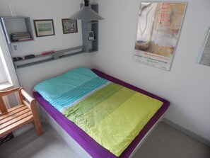 A bedroom with a bed, a chair, a shelf with books and a picture frame, and a poster on the wall.