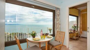 Un balcon avec une table et des chaises, une vue sur la mer et une porte donnant sur une pièce où se trouve un chariot à plantes.