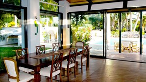 Un comedor con una larga mesa de madera y sillas, ventanas amplias y vistas a una piscina y jardín.