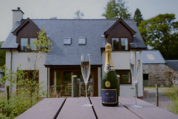 A bottle of champagne and two glasses on a wooden table with a house in the background. - Shepherds Rest self catering sleeps 6, pets welcome. (Fort William)