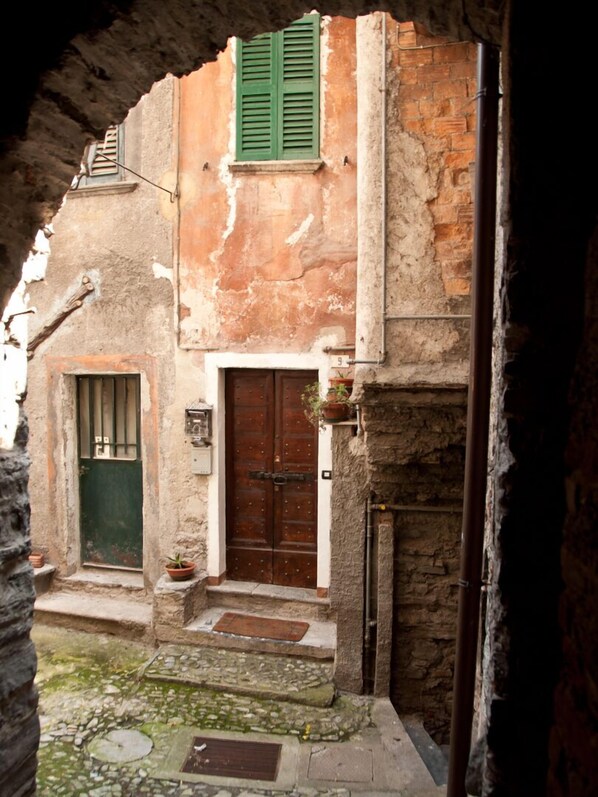 A narrow alleyway with a rustic building featuring a wooden door, a green shuttered window, and a small potted plant on the steps.