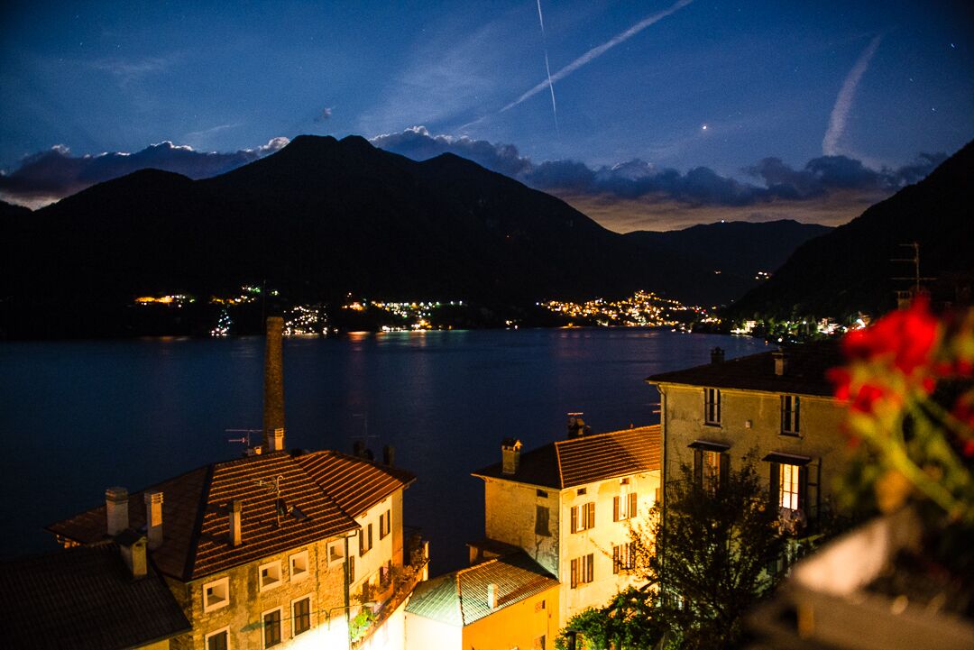 A nighttime view of a lakeside town with buildings along the shore, a mountain range in the background, and a clear sky.