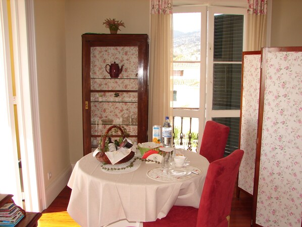 Un comedor con una mesa dispuesta para dos personas, un armario de madera con papel tapiz floral, y vistas al exterior a través de una ventana con contraventanas.