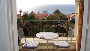 Ein Balkon mit Tisch und zwei Stühlen, mit Blick auf eine Küstenlandschaft mit Gebäuden und Bäumen.