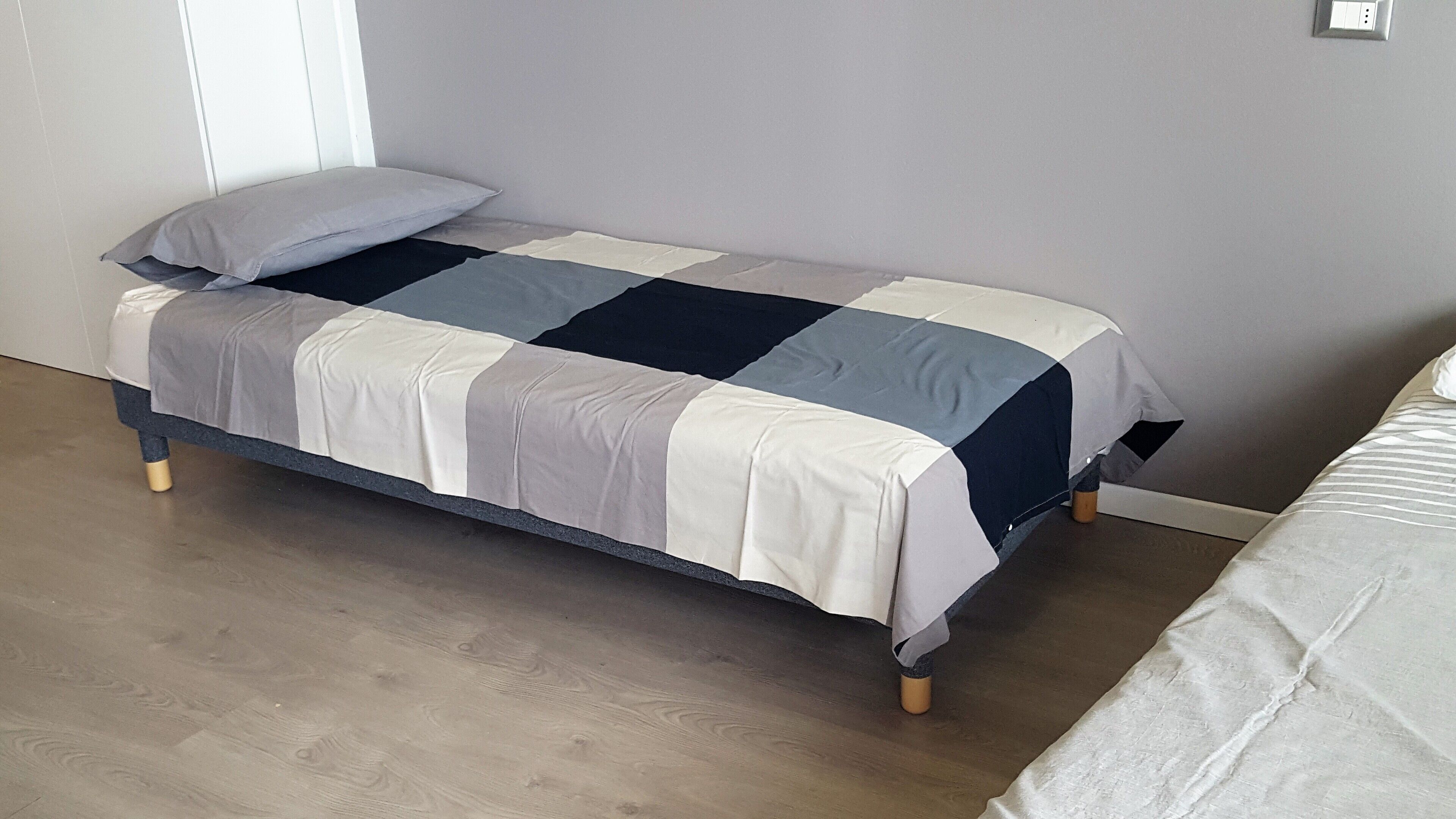 Ein Einzelbett mit karierter Bettdecke in einem Zimmer mit Holzdielenboden und schlichter Wand.