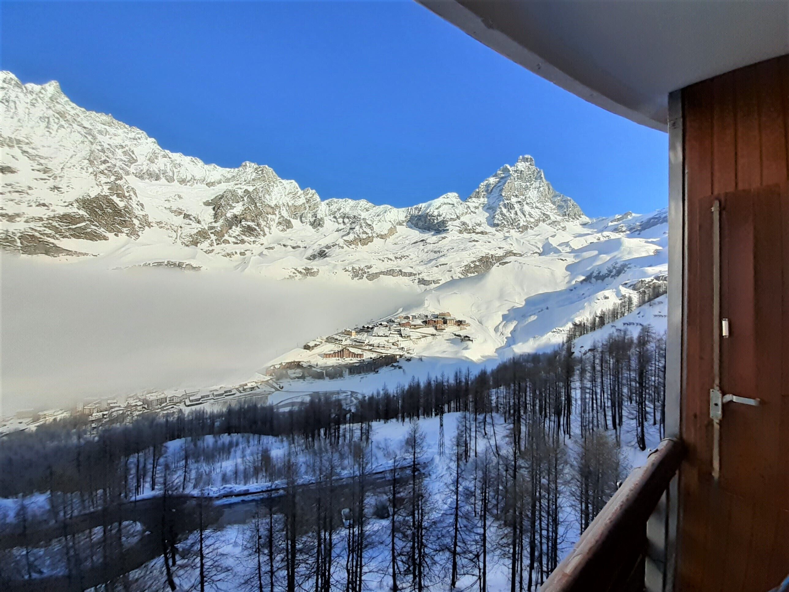 Vista sulle montagne da un balcone, con cime innevate e una valle boscosa sottostante.