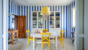A dining area with a white table and yellow chairs, a yellow hanging lamp, and a cabinet with glass doors displaying dishes. - Waterfront villa with panoramic terrace and garden (Fontane Bianche)