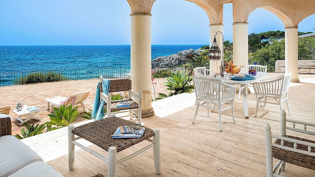 A covered outdoor seating area with a view of the sea, featuring white furniture and a table set with a bowl of fruit and a vase of flowers. - Waterfront villa with panoramic terrace and garden (Fontane Bianche)