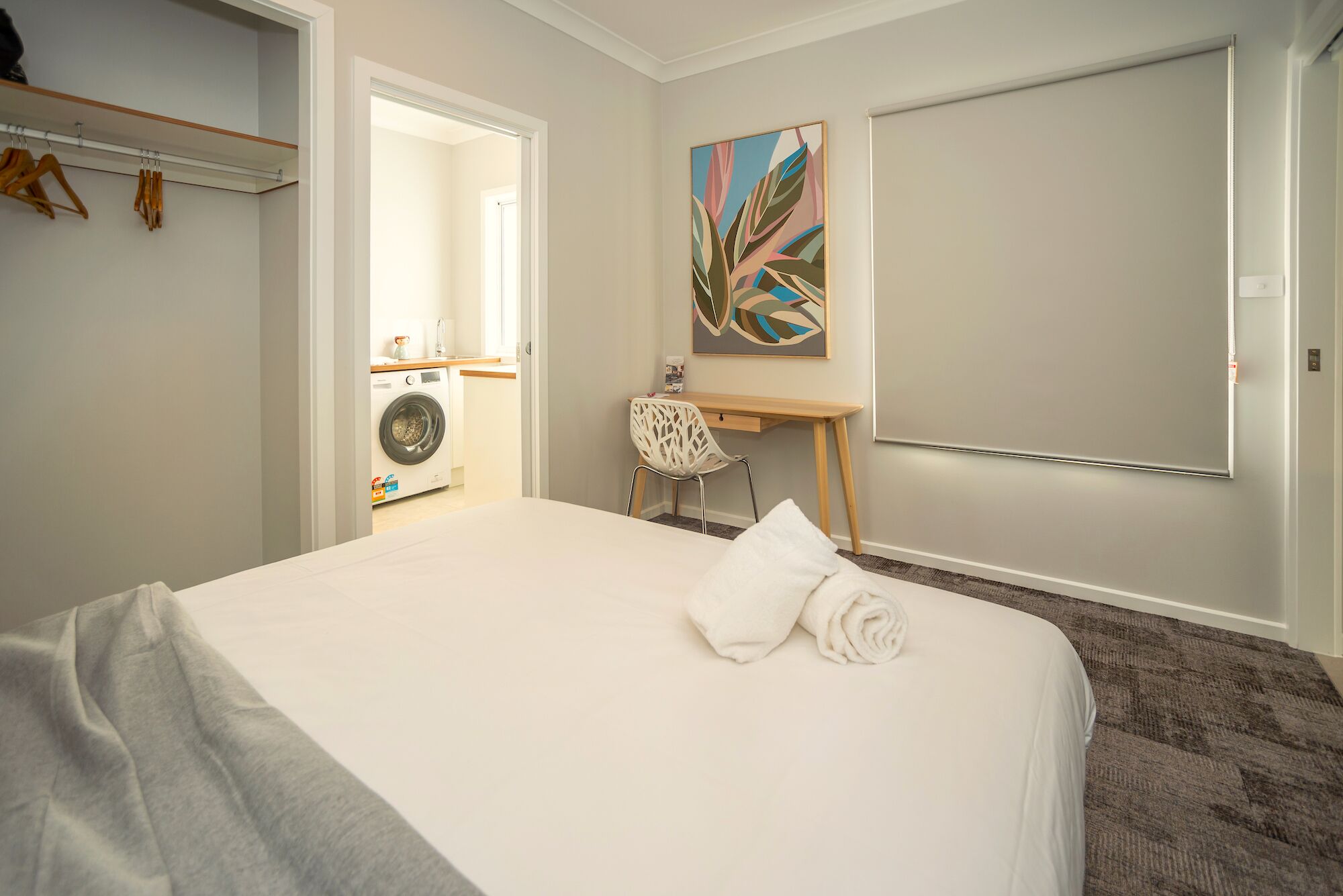 A hotel room with a bed, a desk, a chair, a washing machine, and a painting on the wall.