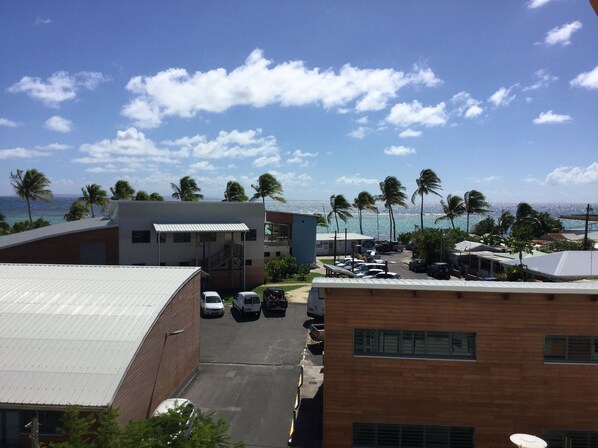 A view of a parking area with several cars, a building with a flat roof, and palm trees in the background.
