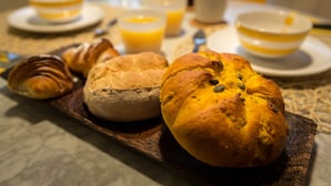 Aamiaispöytä, jossa on croissantteja, leipä ja lasi appelsiinimehua puisella tarjottimella.
