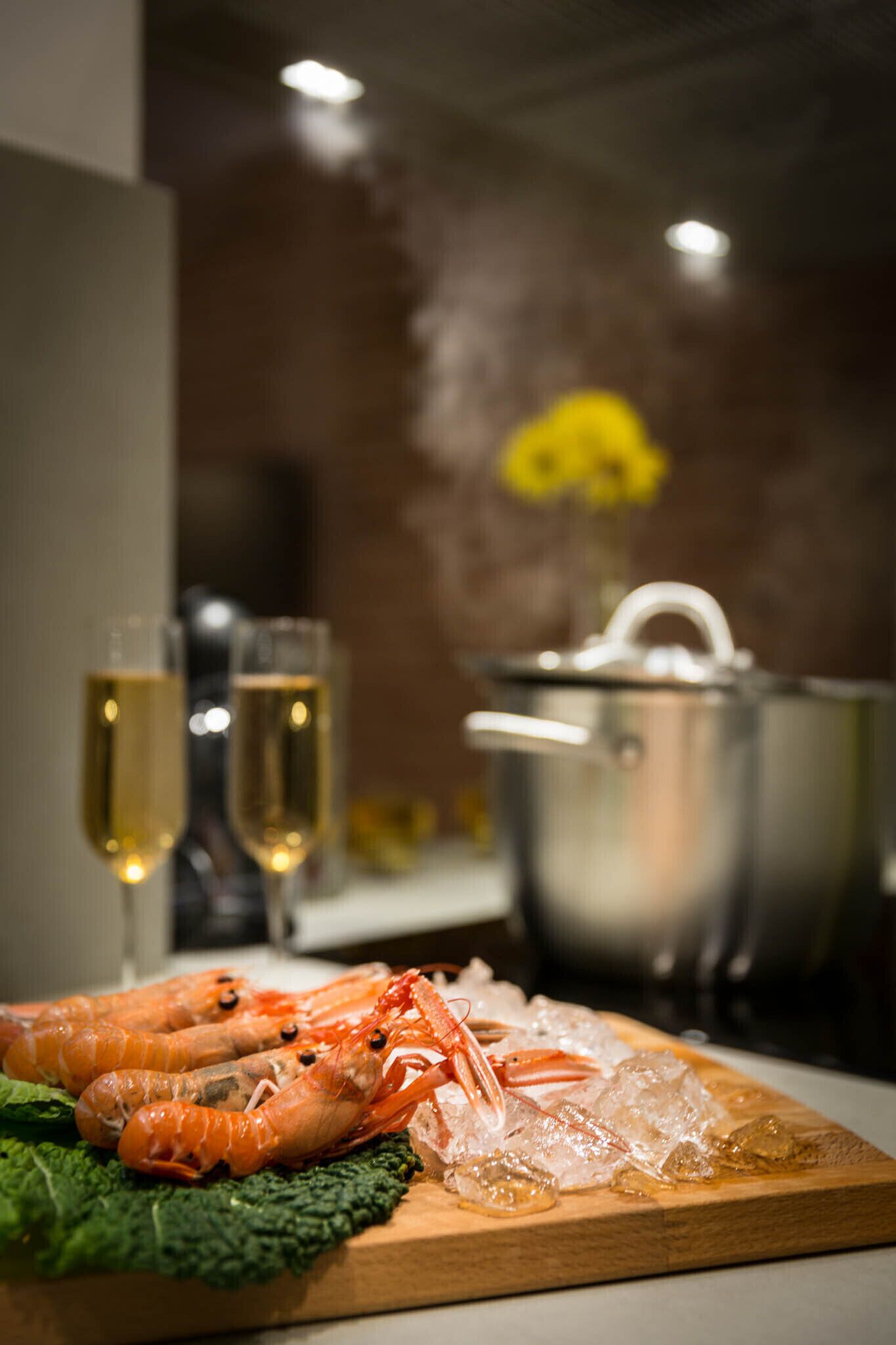 A wooden cutting board with cooked shrimp and ice, a pot, and two glasses of champagne in the background.