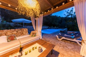 Un área de descanso al aire libre con pared de piedra, una mesa de madera con una botella y copas, un sofá blanco con cojines y una piscina al fondo.