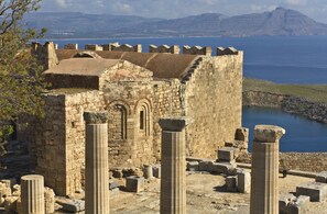Ruines antiques avec des colonnes de pierre, offrant une vue sur la mer et les montagnes en arrière-plan.