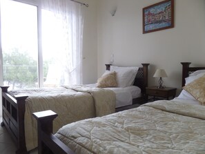 Une chambre avec deux lits, une fenêtre avec des rideaux légers, une table de chevet avec une lampe et un tableau au mur.