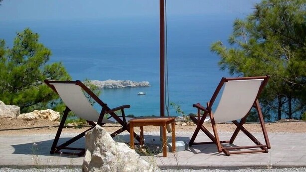 Dues cadires de relax amb una ombria, una taula petita i vista al mar i la costa rocosa.
