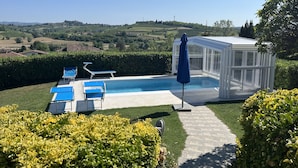 En swimmingpool med liggestole og en tilbygning i glas, omgivet af velplejede hække og fri udsigt over landskabet.