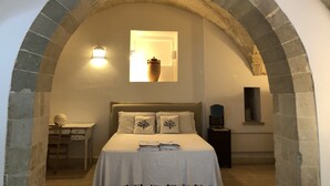 Een slaapkamer met een stenen boog, een bed met een witte spreideken, een nachtkastje, een lamp en een raam met een vaas.