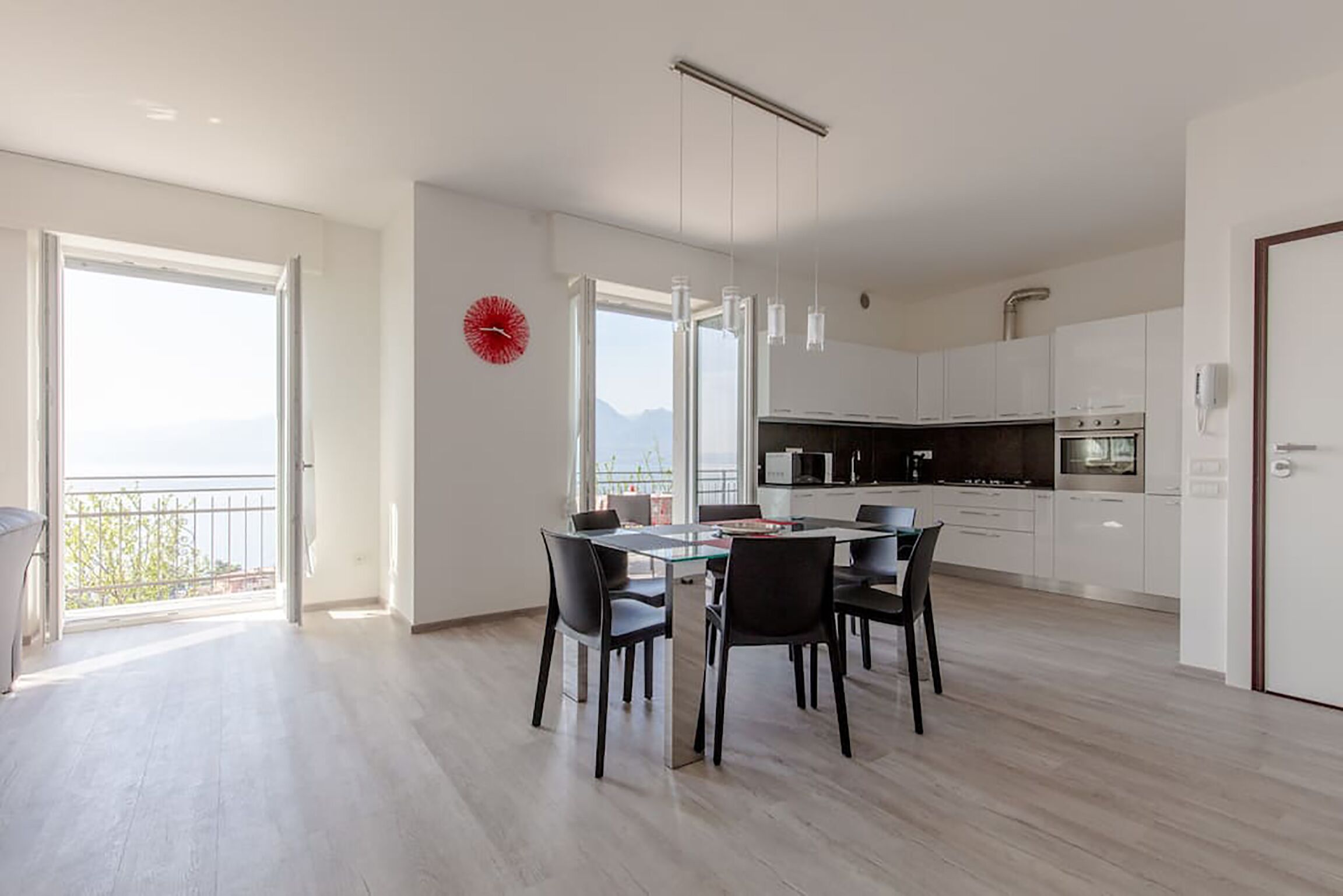 Una cocina moderna con armarios blancos, un comedor con mesa de vidrio y sillas negras, y vistas al exterior a través de amplios ventanales.