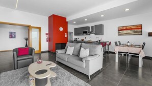 A modern living room with a grey sofa, a red accent wall, a dining area with a table set for four, and a kitchen in the background. - A&M House - Luxury Holidays Home (Arco da Calheta)